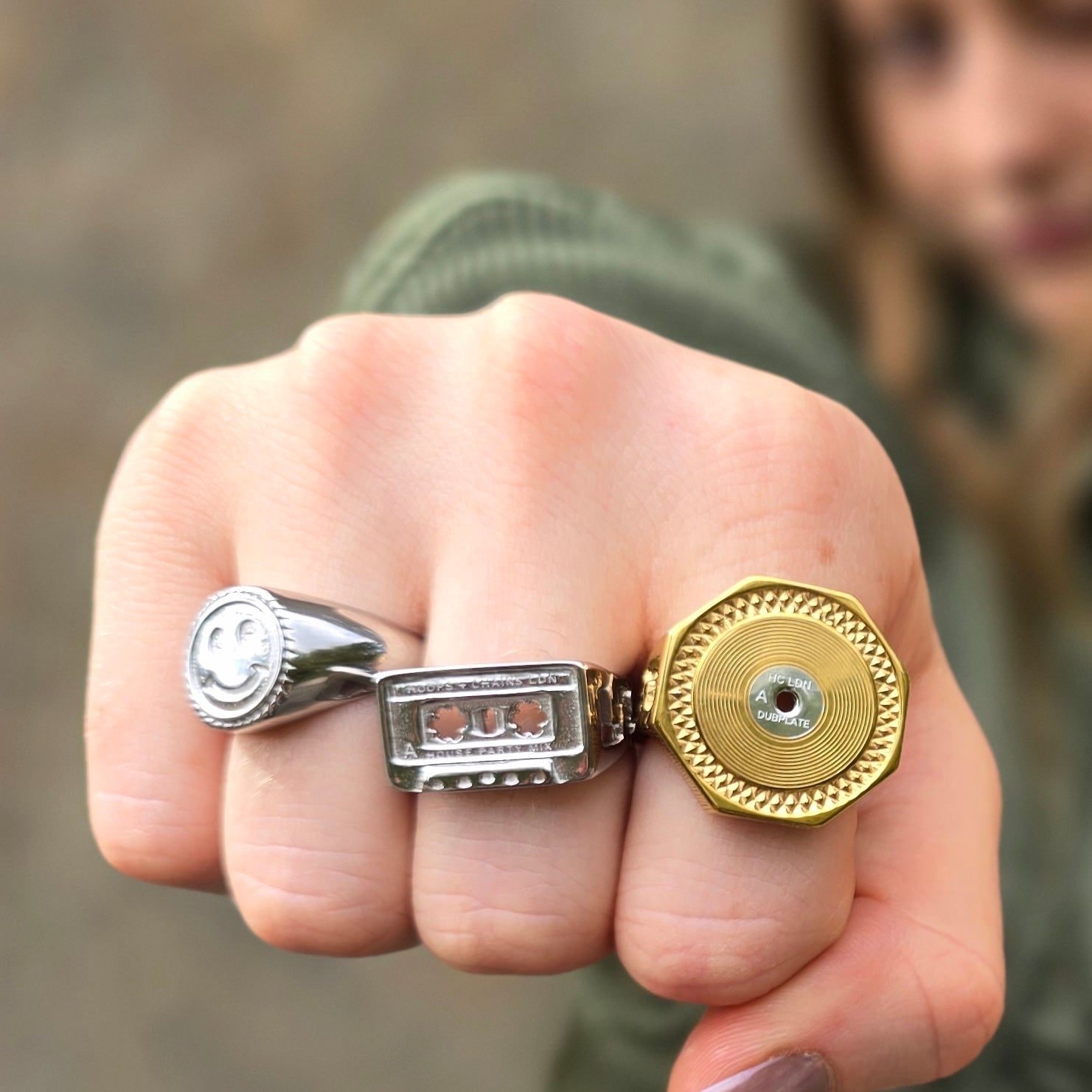 A close-up of a hand wearing three unique rings: a silver ring with a smiley face, a silver ring with a cassette player design, and a gold ring with a textured circular face.