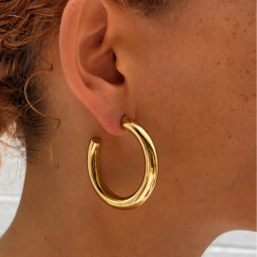 A close-up of a person wearing a thick, polished gold hoop earring on their ear, with curly copper-brown hair and a light pink scrunchie visible.