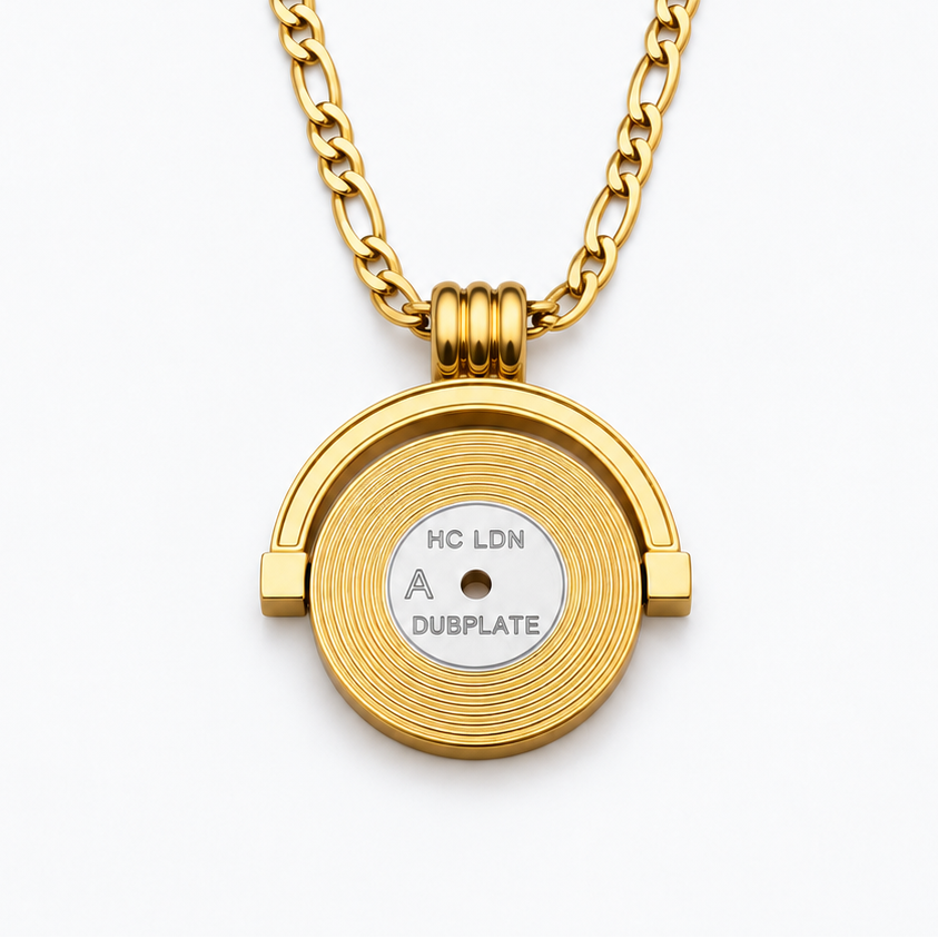 A gold pendant necklace featuring a circular, textured gold disc with a small silver-colored center insert that reads 'HC LDN DUSPLATE'. The necklace is displayed on a clean, light gray background.