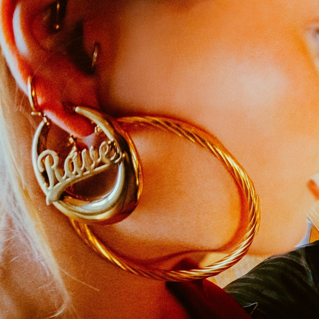 A close-up lifestyle shot of a woman applying lipstick while looking into a small mirror on a wooden table. She is wearing a large gold hoop earring with the name 'Rae' written on it.